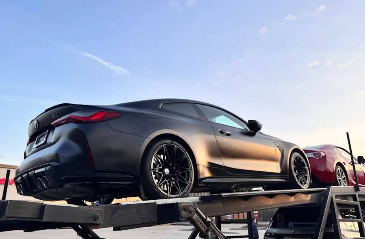 A matte grey BMW M4 loaded on the upper deck of an open car hauler beside a red coupe.