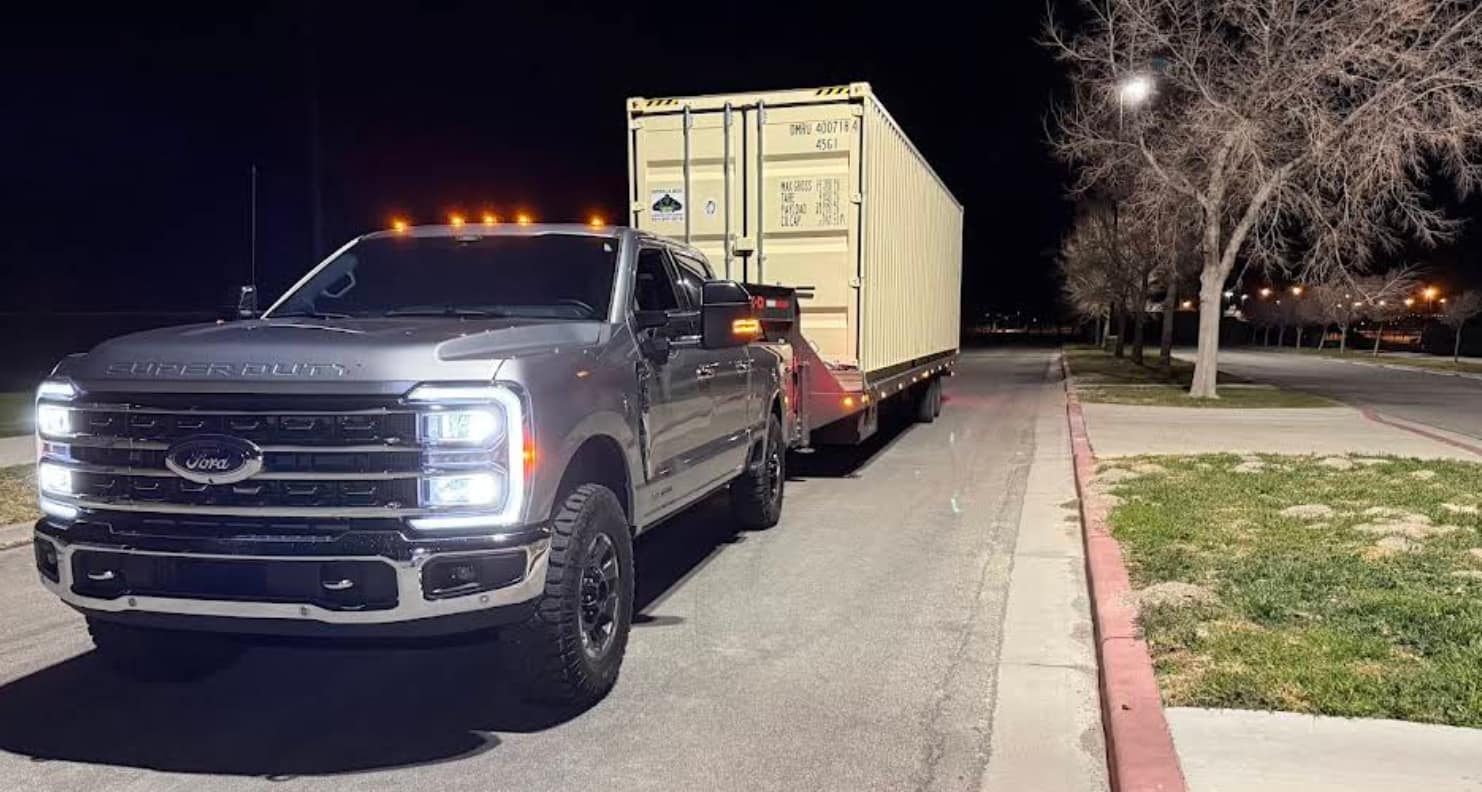 A Ford Super Duty and gooseneck flatbed loaded with a shipping container, parked on a street at night with the truck's lights on.