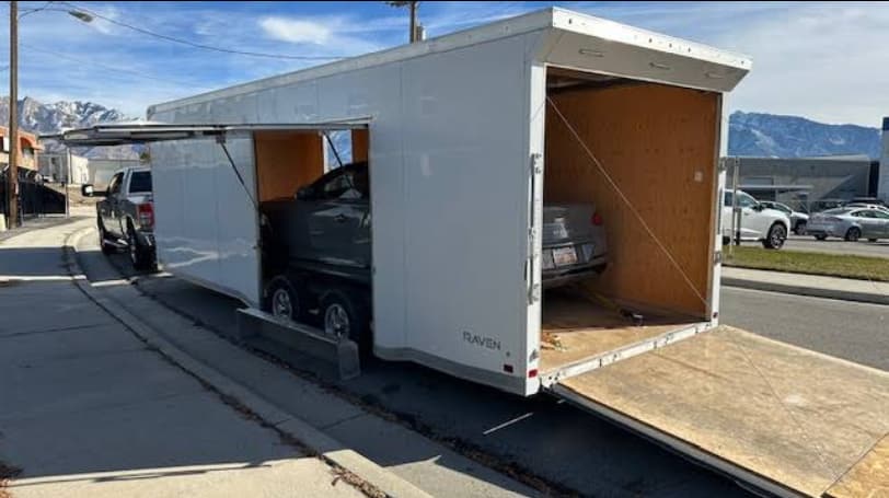 An enclosed car trailer with both doors open and two cars inside, parked on a street with the Wasatch range behind it.