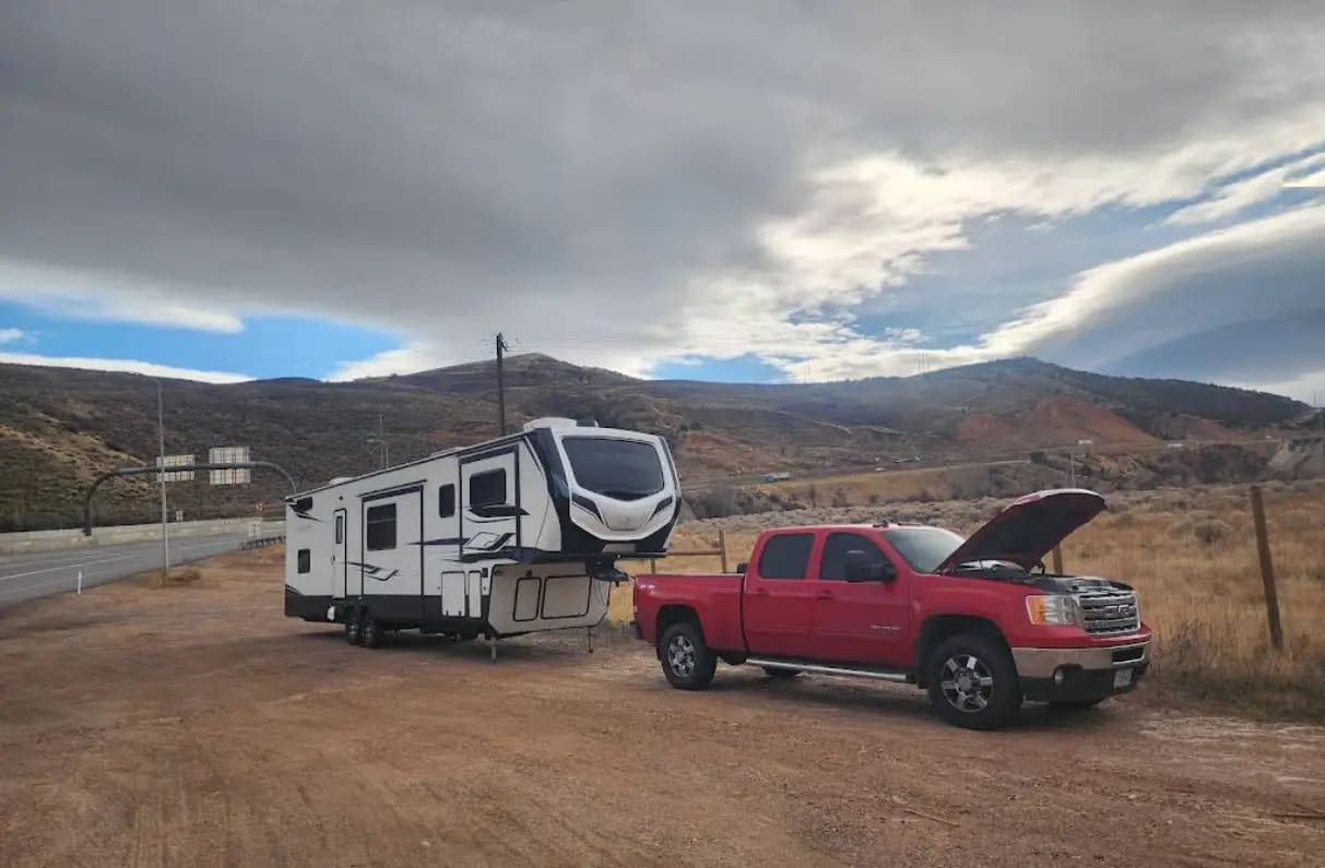 A red GMC pickup hitched to a fifth-wheel trailer on a gravel pull-off beside a highway in dry hill country.