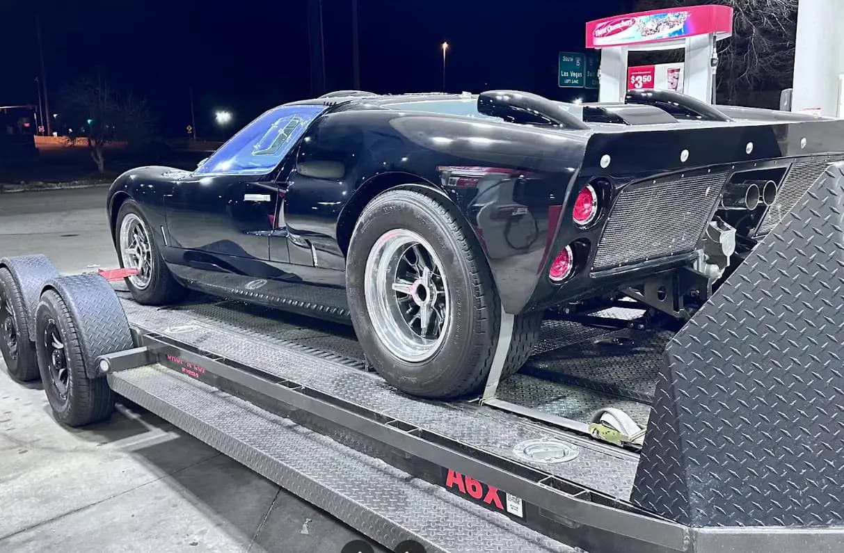 A black GT40 strapped to an open trailer at a fuel stop at night, under a highway sign reading South Las Vegas.