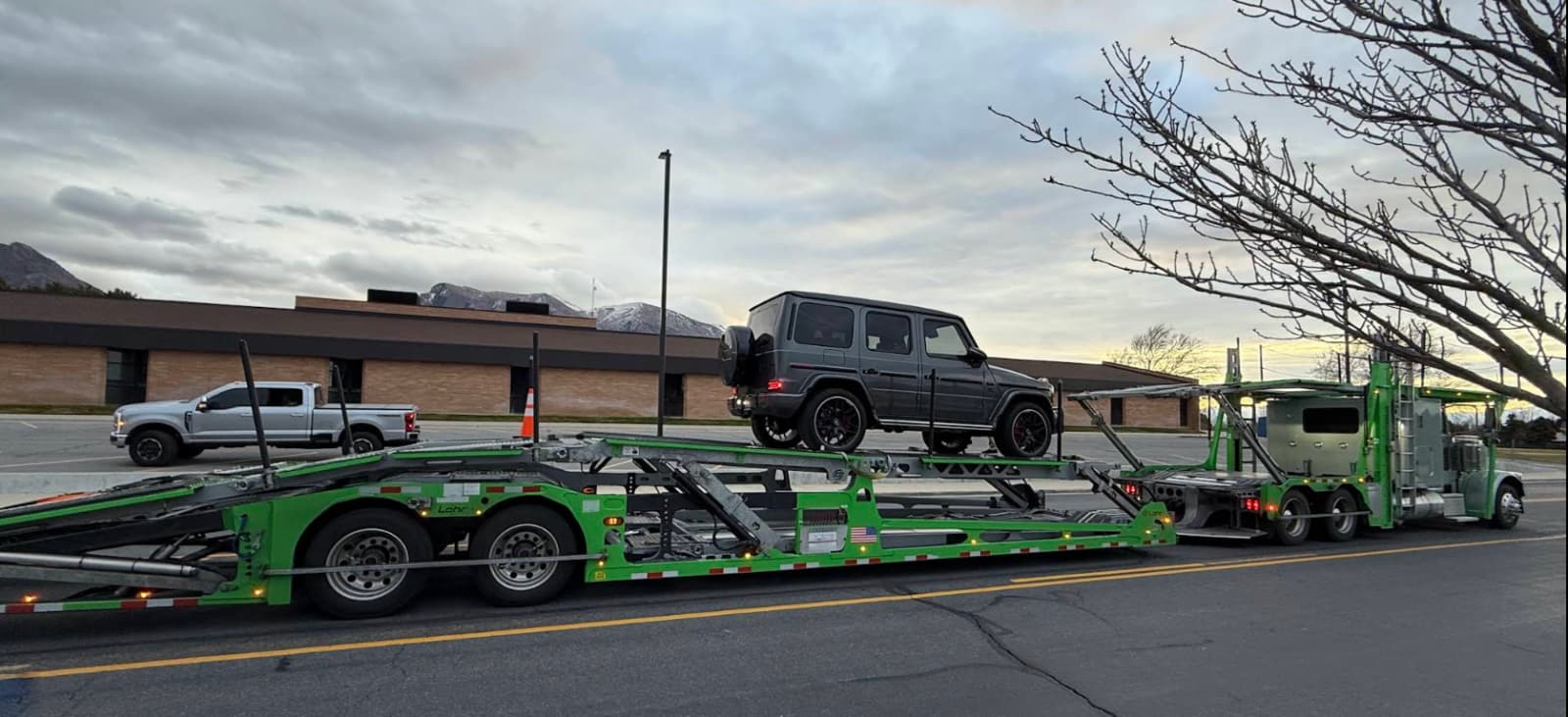 A Mercedes G-Wagon raised on the upper deck of a green multi-car carrier in a parking lot with mountains behind.