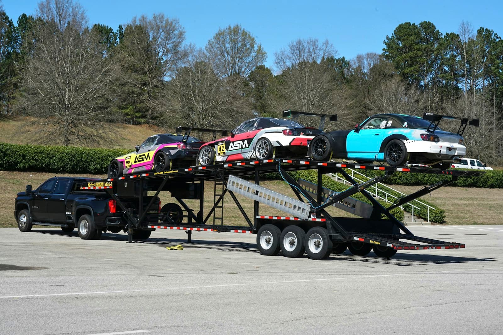 A black dually pickup towing an open multi-car trailer loaded with race cars.